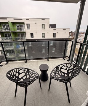 Un balcon avec deux chaises noires et une petite table, donnant sur un paysage urbain avec des bâtiments modernes.