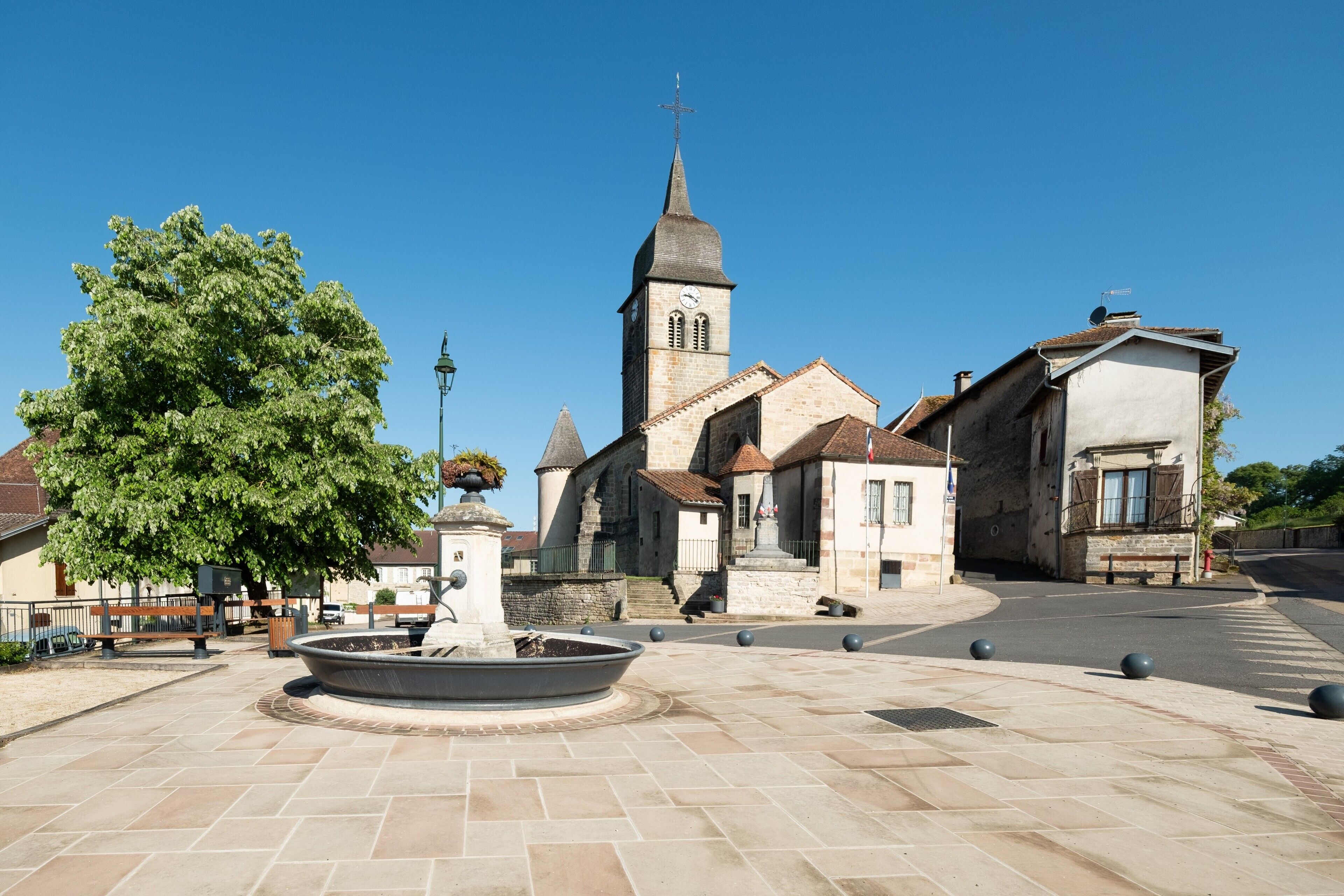 Una iglesia con una torre de reloj, una fuente y una plaza pavimentada con bancos.