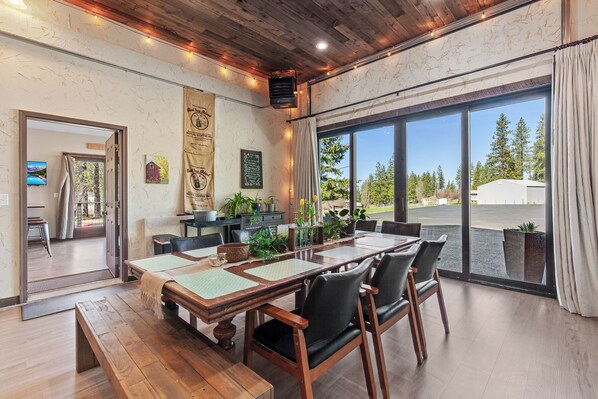 A dining area with a wooden table and chairs, a large window with a view of trees and a building, and a wall with a coffee banner. - New Listing! Large Barn Home | Rustic-Chic Retreat | Hot Tub | Sauna (Coeur d'Alene)