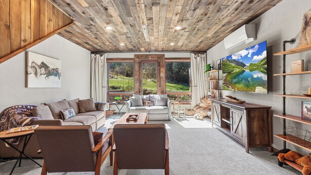 Un salon chaleureux avec un plafond en bois, une grande fenêtre donnant sur un paysage verdoyant, un canapé, une table basse et un téléviseur fixé au mur.