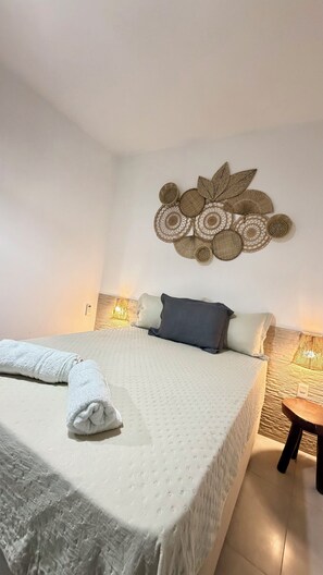 Ein ordentlich gemachtes Bett mit weißer Decke, einem aufgerollten weißen Handtuch und einem dunkelgrauen Kopfkissen.
