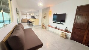 Ein modernes Wohnzimmer mit einer Couch, einem an der Wand befestigten Fernseher, einer Küche im Hintergrund und einem großen Fenster.