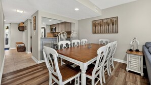 Ein Essbereich mit Holztisch und weißen Stühlen, im Hintergrund eine Küche und ein graues Sofa.