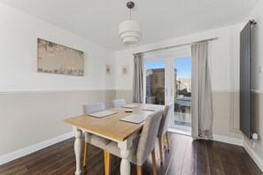 Comedor con mesa de madera, sillas tapizadas en gris, lámpara colgante blanca y vista panorámica de la ciudad a través de la puerta de vidrio.
