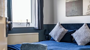 Ein Schlafzimmer mit Bett, Kissen, einem Fenster mit Vorhängen, einem Heizkörper und einer an der Wand befestigten Buddha-Statue.