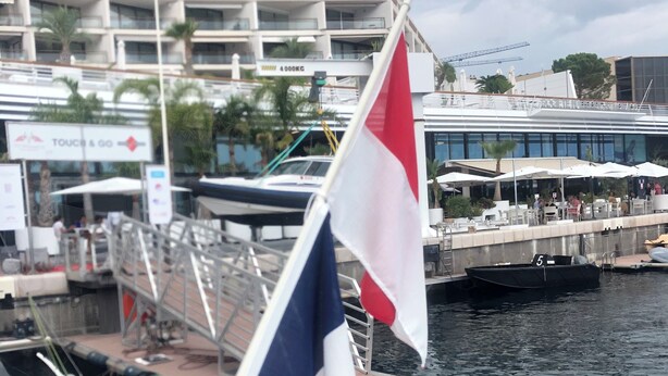 Un puerto deportivo con barcos amarrados, una bandera y un edificio con balcones al fondo.