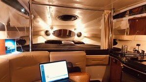 Interior compacto de embarcación con una computadora portátil sobre un asiento acolchado, una pequeña cocina y una cama con cabecera.