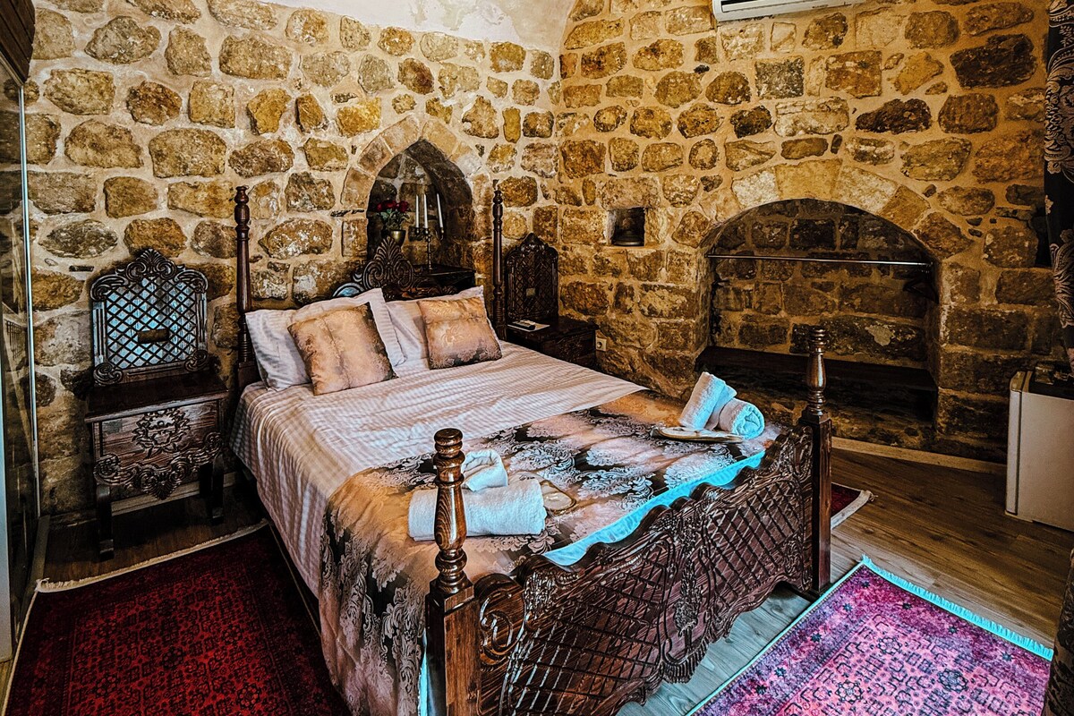 A bedroom with a stone wall, a bed with a wooden headboard, a nightstand, a dresser, and a fireplace.