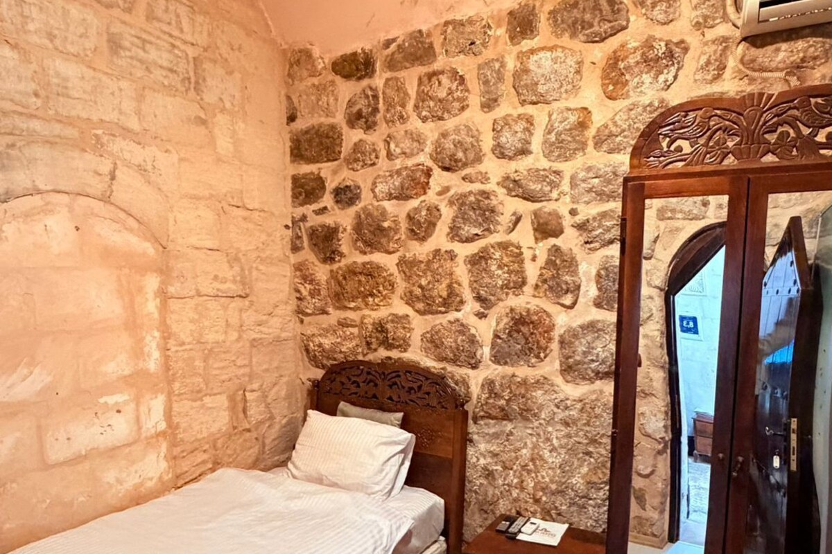 A room with a stone wall, a bed with a wooden headboard, a nightstand, a red patterned rug, and a mirror with a wooden frame.