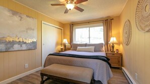 Un dormitorio con cama, banqueta, ventilador de techo, dos lámparas de noche, ventana con cortinas y un cuadro en la pared.