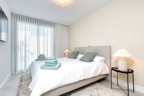 Ein modernes Schlafzimmer mit einem großen Bett, zwei Nachttischlampen, einem Nachttisch und Blick ins Freie durch durchscheinende Vorhänge.