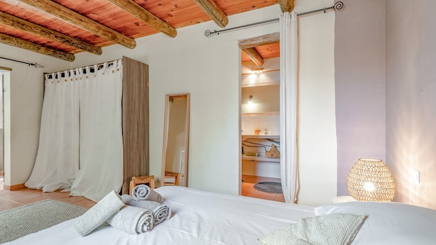 Un dormitorio con techo de madera, cama con ropa de cama blanca, lámpara de fibras naturales y vista al baño.