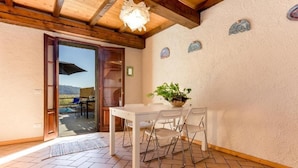 A room with a white table and chairs, a potted plant, a wooden ceiling, and a door leading to an outdoor area with a pool and chairs. - Casa dei Ciliegi (Bagni di Lucca)