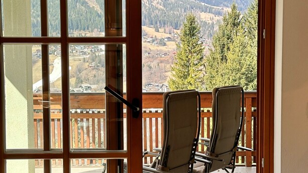Un'area pranzo con tavolo apparecchiato per due, vista su un paesaggio montuoso attraverso una grande finestra e un terrazzo in legno esterno.