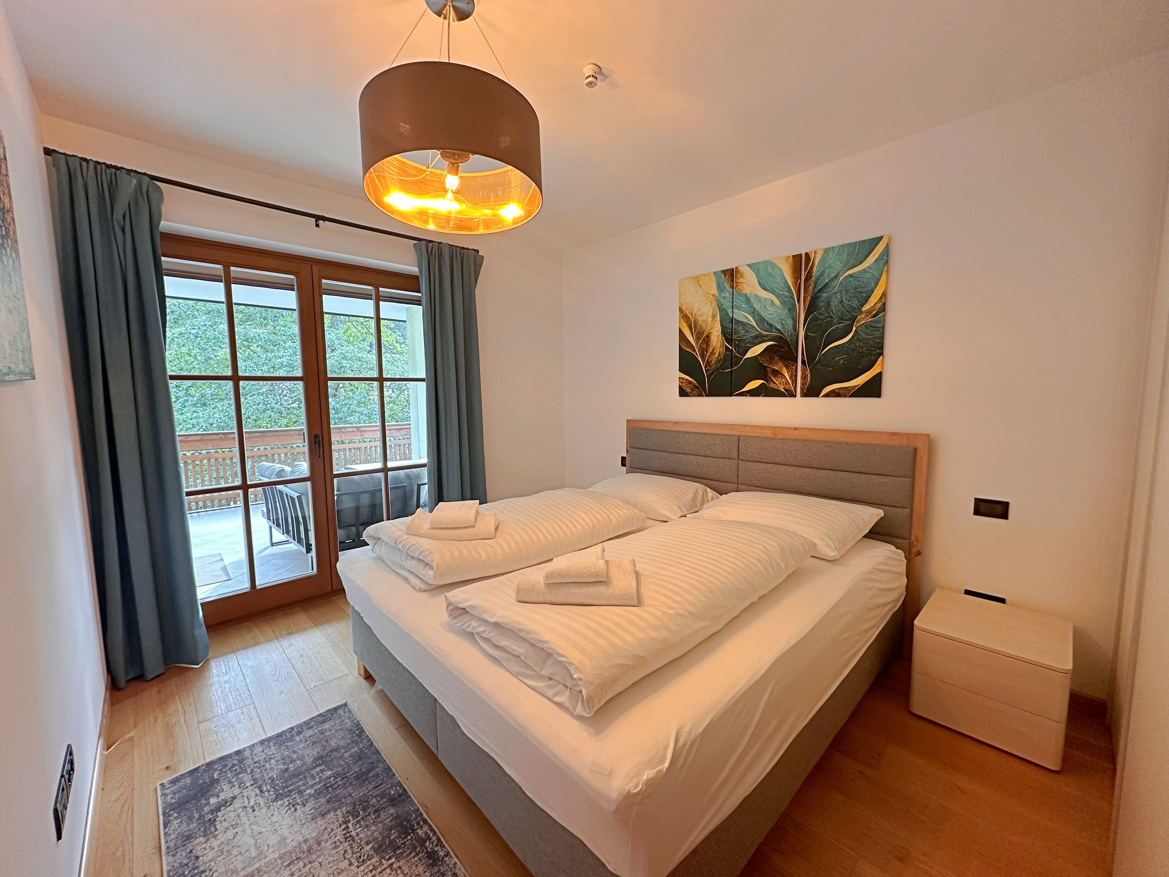 Ein Schlafzimmer mit einem Bett, einem Nachttisch, einem Fenster mit Vorhängen, einem Gemälde an der Wand und einer Deckenleuchte.