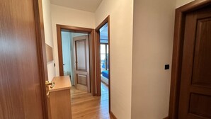 Un corridoio con pavimento in legno, una porta in legno sulla destra e vista su un balcone con ringhiera blu.