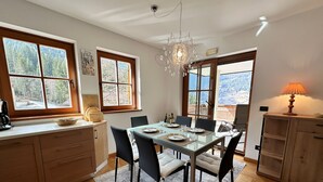 Zona pranzo con tavolo in vetro, sedie, un lampadario, pensili in legno e finestre con vista su alberi e montagne.