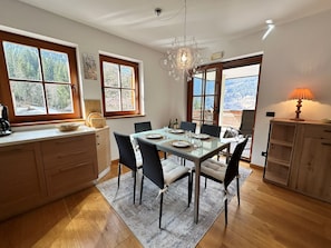 A dining area with a glass table, chairs, a chandelier, wooden cabinets, and windows with a view of trees and mountains. - ALPINE VIEW Apartment Bad Kleinkirchheim (Bach)