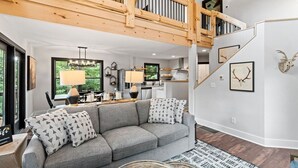 Un amplio salón con un sofá gris, una escalera de madera y vistas al exterior.