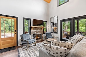 A spacious living room with a stone fireplace, comfortable seating, and large windows offering a view of the outdoors. - Deer Creek Cabin | Hot Tub & Fire Pit (Hendersonville)