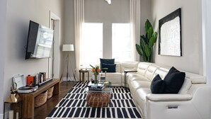 A living room with a sofa, a coffee table, a floor lamp, a TV, and a large plant. - New 2-bedroom corporate loft in charming Atlanta, right by the airport. (Atlanta)