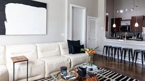 A living room with a sofa, a coffee table, and a kitchen area with stools. - New 2-bedroom corporate loft in charming Atlanta, right by the airport. (Atlanta)