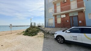 Un bâtiment devant lequel est garée une voiture blanche, une plage en arrière-plan et un ciel dégagé.
