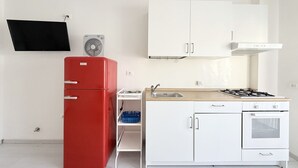 Une cuisine de taille réduite avec des meubles blancs, un réfrigérateur rouge, une cuisinière à gaz et un ventilateur fixé au mur.