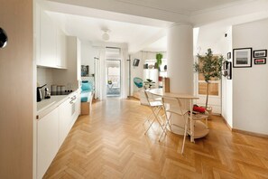 A modern kitchen with white cabinetry, a dining area with a round table and chairs, and a living space with a sofa and a TV. - Estia Premium Apartments - Capri Blue Salonica (Thessaloniki)