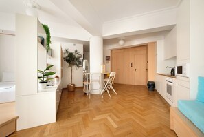 Un apartamento moderno con suelo de madera en diseño de espiga, zona de comedor con mesa y sillas blancas, cocina con armarios blancos y sofá azul.
