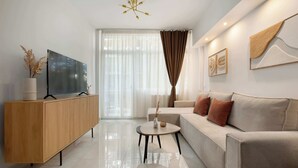 A modern living room with a sofa, a TV on a wooden cabinet, a round coffee table, and a large window with curtains. - Estia Premium Apartments - Superior Suite Vackhou 1 #1.2 (Thessaloniki)