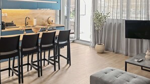 Une cuisine moderne avec des meubles bleus, un coin repas avec des chaises noires, un canapé gris et un téléviseur posé sur une table basse.