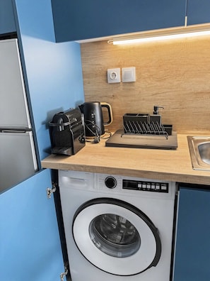 A compact kitchen with a washing machine, a coffee maker, a kettle, and a soap dispenser on the countertop. - Estia Premium Apartments - Valouritou 52 Sqm2 Apartment (Thessaloniki)
