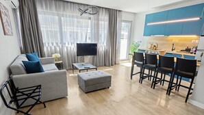 Ein modernes Wohnzimmer mit Sofa, Ottoman und Fernseher. Es gibt einen Essbereich mit Stühlen und eine Küche mit blauen Schränken.