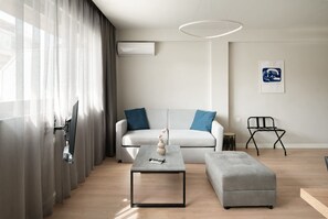 Ein modernes Wohnzimmer mit einem grauen Sofa, einem Couchtisch und einem grauen Hocker. Ein schwarzer Sessel und ein kleiner runder Tisch mit einem dekorativen Gegenstand auf dem Couchtisch sind ebenfalls zu sehen.