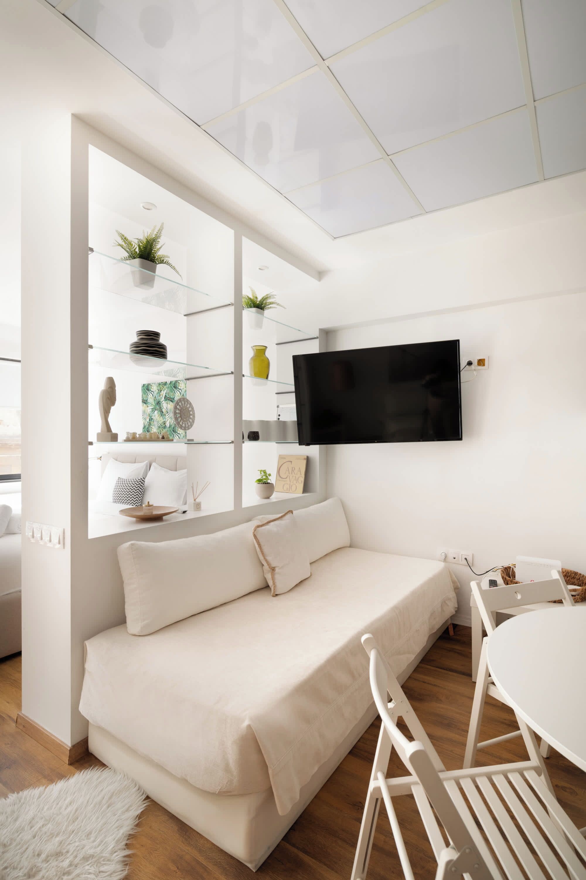 Un salon moderne avec un canapé blanc, un téléviseur fixé au mur et un coin repas comprenant une table et des chaises blanches.
