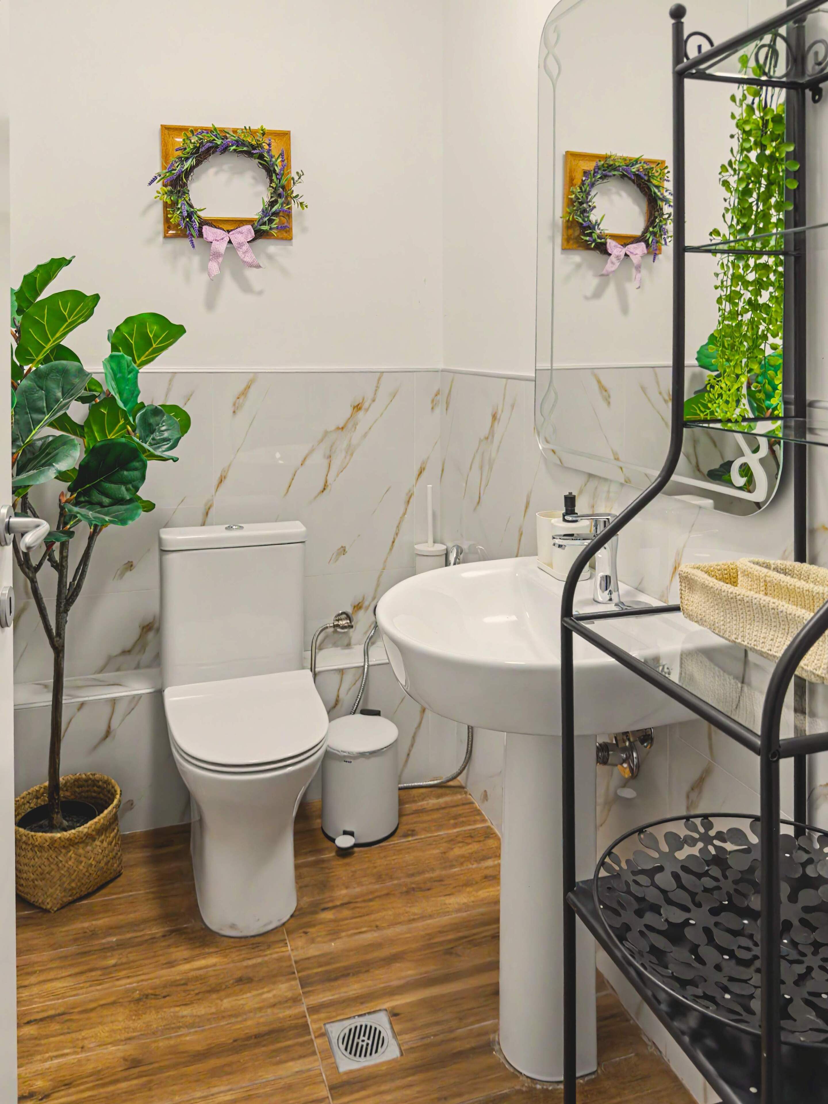 A bathroom with a toilet, sink, and mirror, with two wreaths on the wall and a plant on the left.