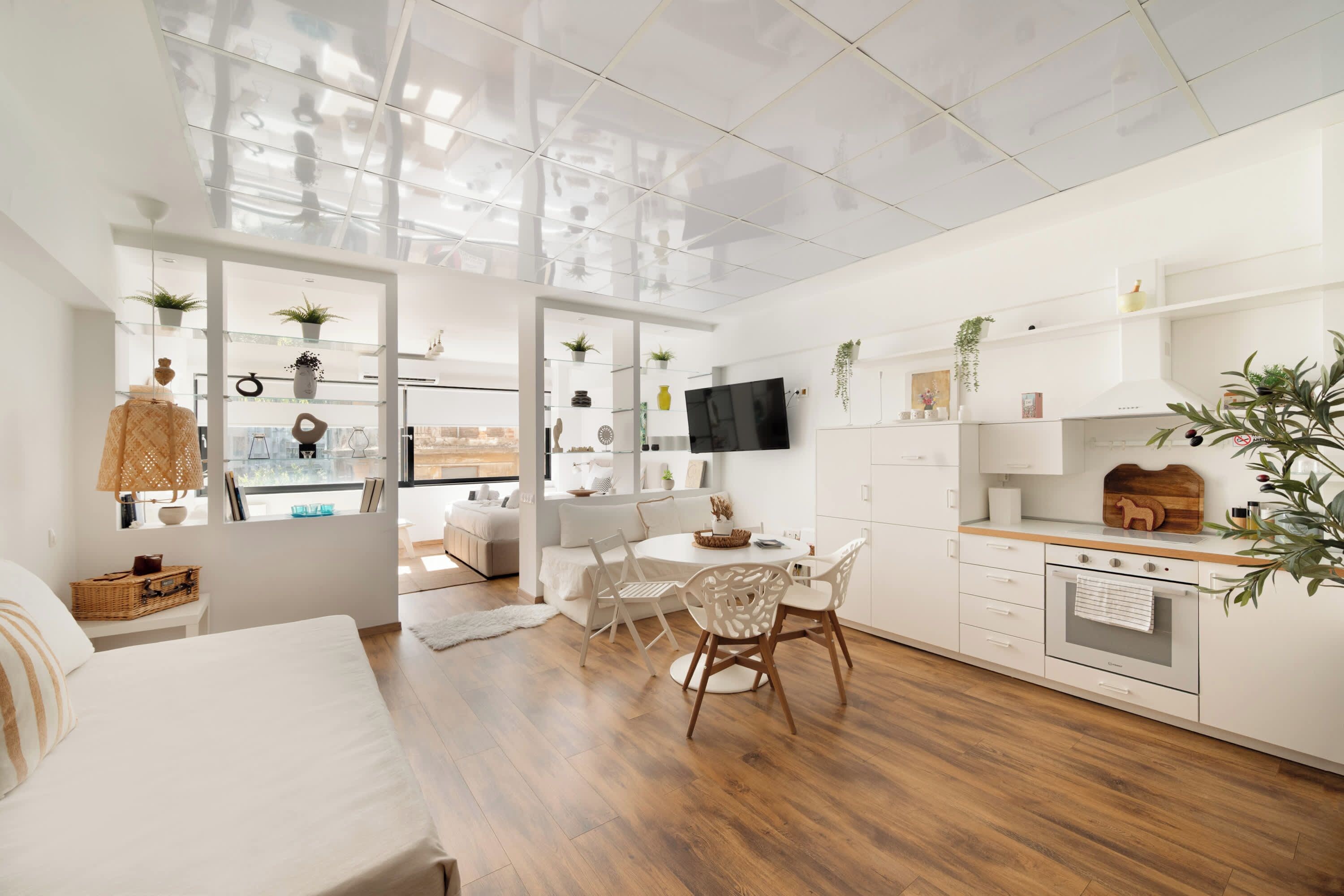 Quarto moderno e bem iluminado, com piso de madeira, área de jantar com mesa e cadeiras brancas, e cozinha com armários brancos e uma TV fixada na parede.