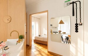 A modern hotel room with a wooden floor, a white bed, a kitchen area with a stove and bar stools, and a bathroom with a gold faucet. - Estia Premium Apartments - Honey Paleon Patron Germanou (Thessaloniki)