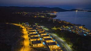 Un hotel costero con hileras de habitaciones uniformemente iluminadas, una carretera y vistas al mar y luces de la ciudad a lo lejos.