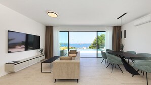 Una sala de estar moderna con un televisor de pantalla plana, un sofá, una mesa de comedor con sillas y un amplio ventanal corredera que da a un balcón con vistas al mar.