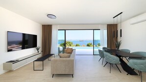Uma sala de estar moderna com televisão de tela plana, sofá, mesa de jantar com cadeiras e uma grande janela com vista para o mar.