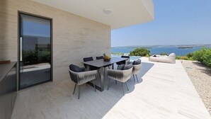 Uma área moderna para refeições ao ar livre, com mesa, cadeiras, porta de vidro e vista para o mar.
