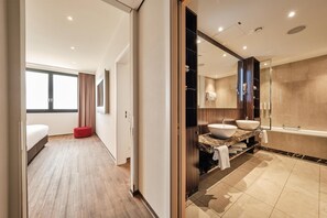 Una habitación de hotel moderna con un ventanal grande, un reposapiés rojo, un baño con bañera y una encimera de mármol.