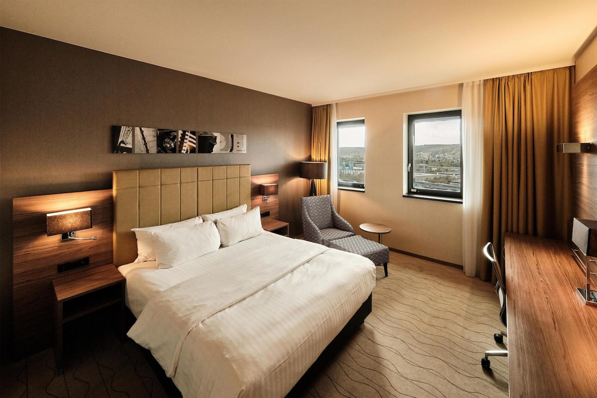 A hotel room with a large bed, a desk, a chair, a small table, and a view of the city.