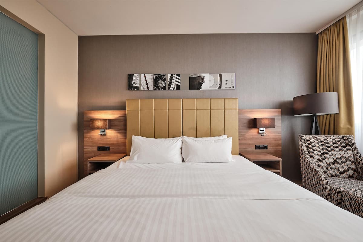 A hotel room with a large bed, a headboard with a panel design, two bedside tables with lamps, a patterned armchair, and a wall-mounted artwork.