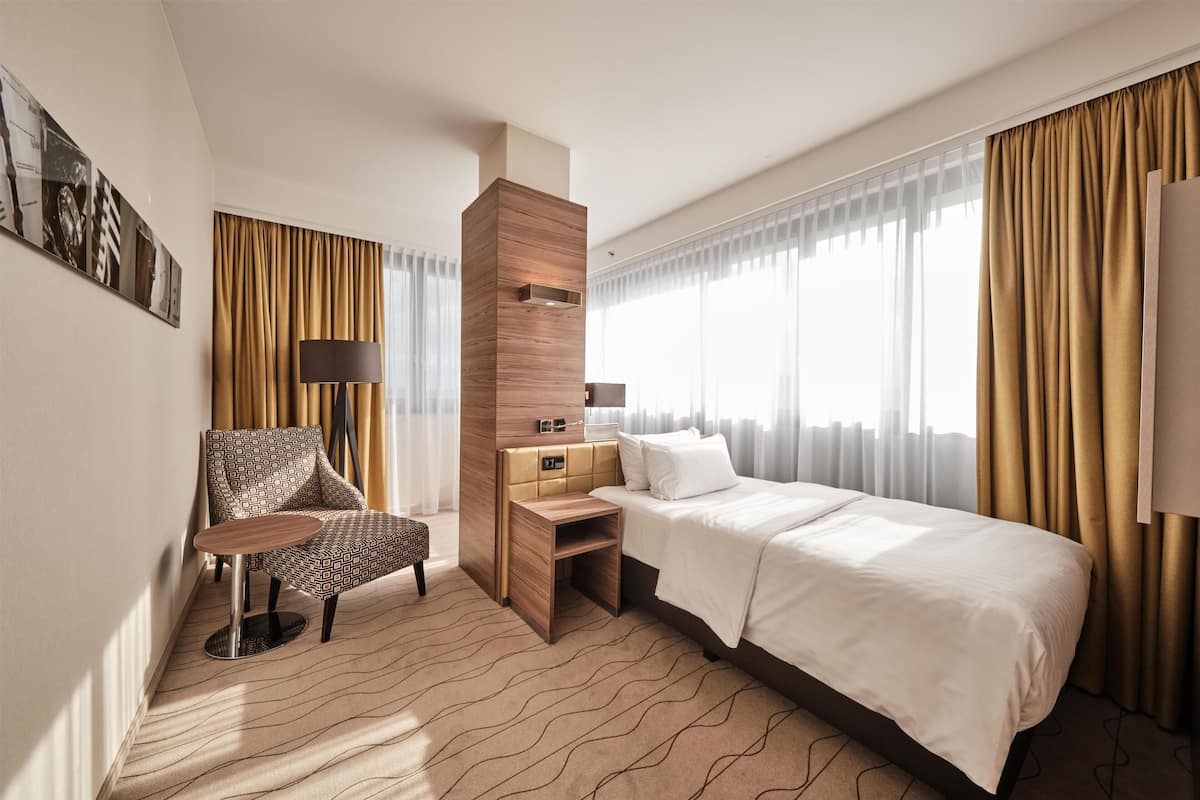 A hotel room with a bed, a desk, a chair, a lamp, and a large window with curtains.