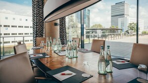 A conference room with a long table, chairs, water bottles, and a view of buildings through large windows. - Radisson Blu Hotel, Bonn (T) (Bonn)
