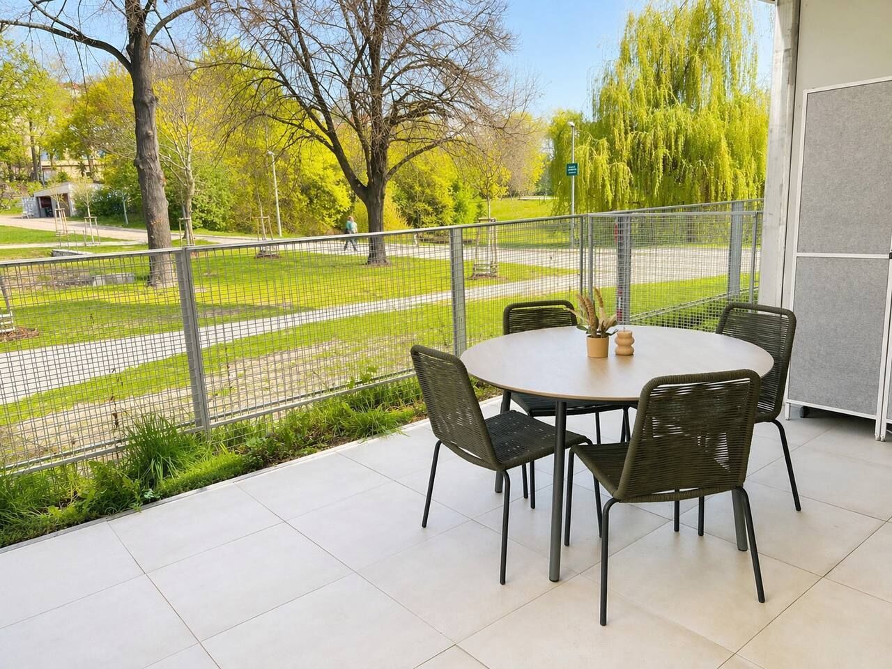 Eine Terrasse mit einem runden Tisch und Stühlen, Blick auf einen Park mit Bäumen und einem Zaun.