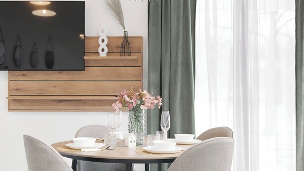 Un comedor con una mesa redonda, sillas, un jarrón con flores, un televisor montado en la pared y un panel de madera.